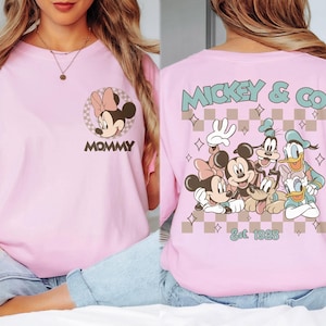 K&ouml;nnte beinhalten: Hellrosa T-Shirt mit Minnie Mouse und dem Wort "MOMMY" auf der Vorderseite. Auf der R&uuml;ckseite ist Mickey & Co. mit Mickey, Minnie, Goofy, Donald und Pluto sowie der Text "Est. 1928" abgebildet.