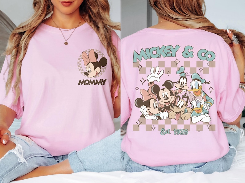 K&ouml;nnte beinhalten: Hellrosa T-Shirt mit Minnie Mouse und dem Wort "MOMMY" auf der Vorderseite. Auf der R&uuml;ckseite ist Mickey & Co. mit Mickey, Minnie, Goofy, Donald und Pluto sowie der Text "Est. 1928" abgebildet.