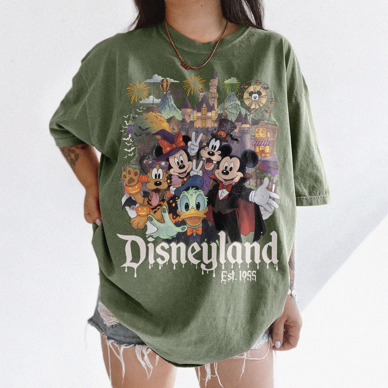 May include: An olive green t-shirt featuring a graphic print of Disney characters in Halloween costumes, including Mickey Mouse, Minnie Mouse, Donald Duck, and Pluto. The design includes a castle, fireworks, and the text Disneyland Est. 1955.