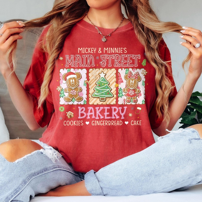 Op de afbeelding: Rood t-shirt met een Main Street Bakery-ontwerp met Mickey en Minnie Mouse peperkoekpersonages en een kerstboom. De tekst bevat "Cookies, Gingerbread, Cake."