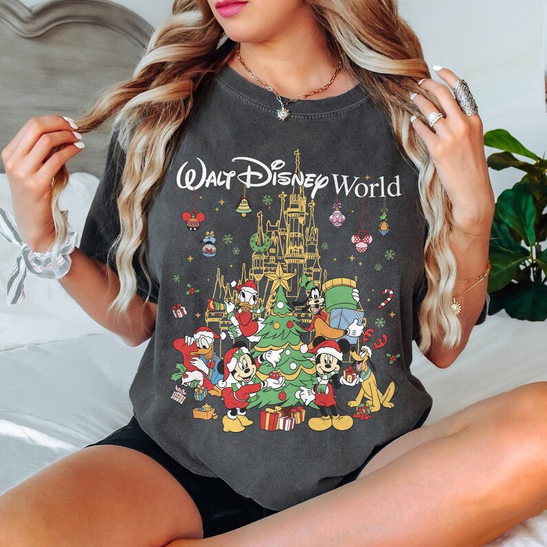 May include: Dark gray t-shirt featuring a festive Walt Disney World design. The graphic includes a castle, Christmas tree, and various Disney characters in holiday attire. The text "Walt Disney World" is arched above the illustration.