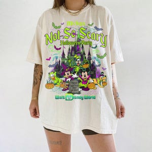 May include: An off-white short-sleeve t-shirt featuring a graphic print of Mickey's Not-So-Scary Halloween Party at Walt Disney World. The design includes Disney characters in costumes, a castle, pumpkins, bats, and spiderwebs in neon green and purple.