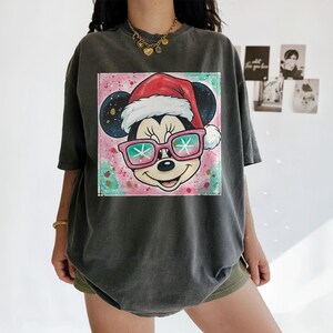 May include: Dark gray t-shirt featuring a colorful graphic of Mickey Mouse wearing a Santa hat and sunglasses with snowflake lenses. The graphic is set against a pink and teal background. The shirt has short sleeves and a relaxed fit.