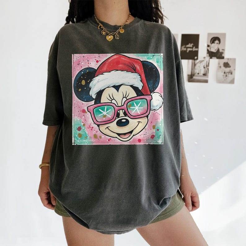 May include: Dark gray t-shirt featuring a colorful graphic of Mickey Mouse wearing a Santa hat and sunglasses with snowflake lenses. The graphic is set against a pink and teal background. The shirt has short sleeves and a relaxed fit.