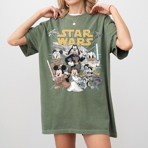 May include: Olive green t-shirt featuring a graphic of Disney characters in Star Wars attire. The Star Wars logo is in yellow. Characters include Mickey Mouse, Minnie Mouse, Donald Duck, and Goofy.