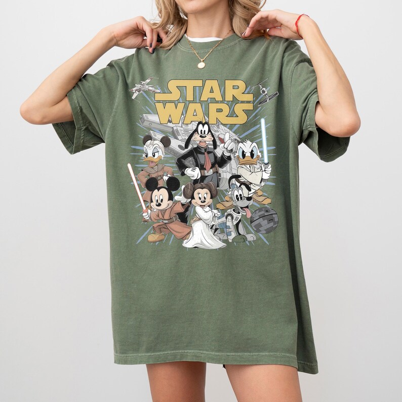 May include: Olive green t-shirt featuring a graphic of Disney characters in Star Wars attire. The Star Wars logo is in yellow. Characters include Mickey Mouse, Minnie Mouse, Donald Duck, and Goofy.