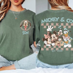 K&ouml;nnte beinhalten: Gr&uuml;nes T-Shirt mit Pluto und dem Wort "SON" in einem Kreis auf der Vorderseite. Auf der R&uuml;ckseite sind Micky Maus, Minnie Maus, Goofy, Donald Duck und Daisy Duck mit "Mickey & Co" und "Est. 1928" abgebildet.