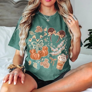 May include: A dark green short-sleeve t-shirt featuring a graphic print of orange pumpkins, mouse ears, a coffee cup, and a string of lights with the text 'TIS THE SEASON'. The design includes autumn leaves and bows in a warm, muted color palette.