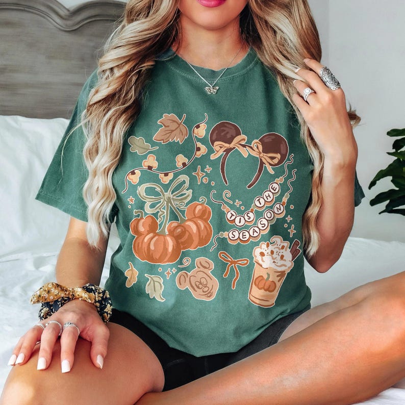 May include: A dark green short-sleeve t-shirt featuring a graphic print of orange pumpkins, mouse ears, a coffee cup, and a string of lights with the text 'TIS THE SEASON'. The design includes autumn leaves and bows in a warm, muted color palette.
