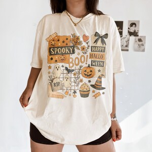 K&ouml;nnte beinhalten: Cremefarbenes T-Shirt zum Thema Halloween mit einem grafischen Design mit K&uuml;rbissen, Geistern, einem Hexenhut und den Worten "Spooky Boo! Happy Halloween". Das Design umfasst Sterne, Blumen und einen Cupcake.
