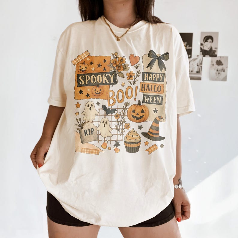 K&ouml;nnte beinhalten: Cremefarbenes T-Shirt zum Thema Halloween mit einem grafischen Design mit K&uuml;rbissen, Geistern, einem Hexenhut und den Worten "Spooky Boo! Happy Halloween". Das Design umfasst Sterne, Blumen und einen Cupcake.