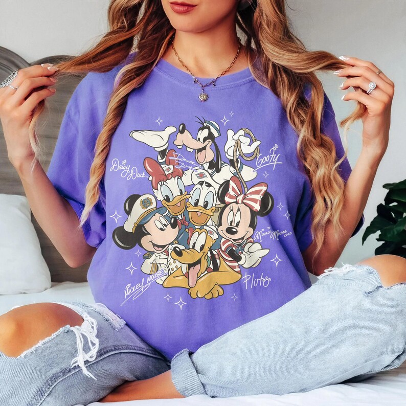 May include: A purple t-shirt featuring a graphic of Disney characters including Mickey Mouse, Minnie Mouse, Donald Duck, Daisy Duck, Goofy, and Pluto. The names of the characters are written in script. The shirt is a solid color.