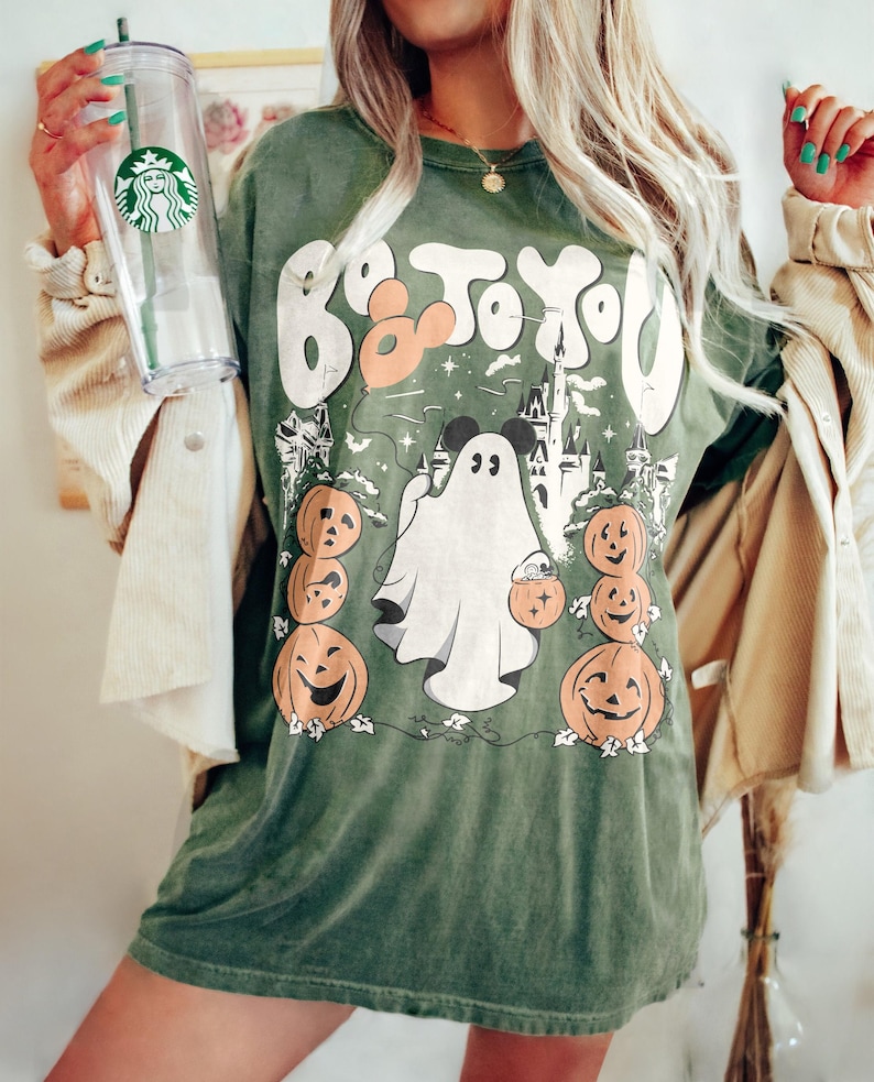 May include: Green t-shirt with a Halloween graphic. The design includes a ghost, pumpkins, and a castle, with the text "Boo To You" in white and orange. The shirt has a loose fit.