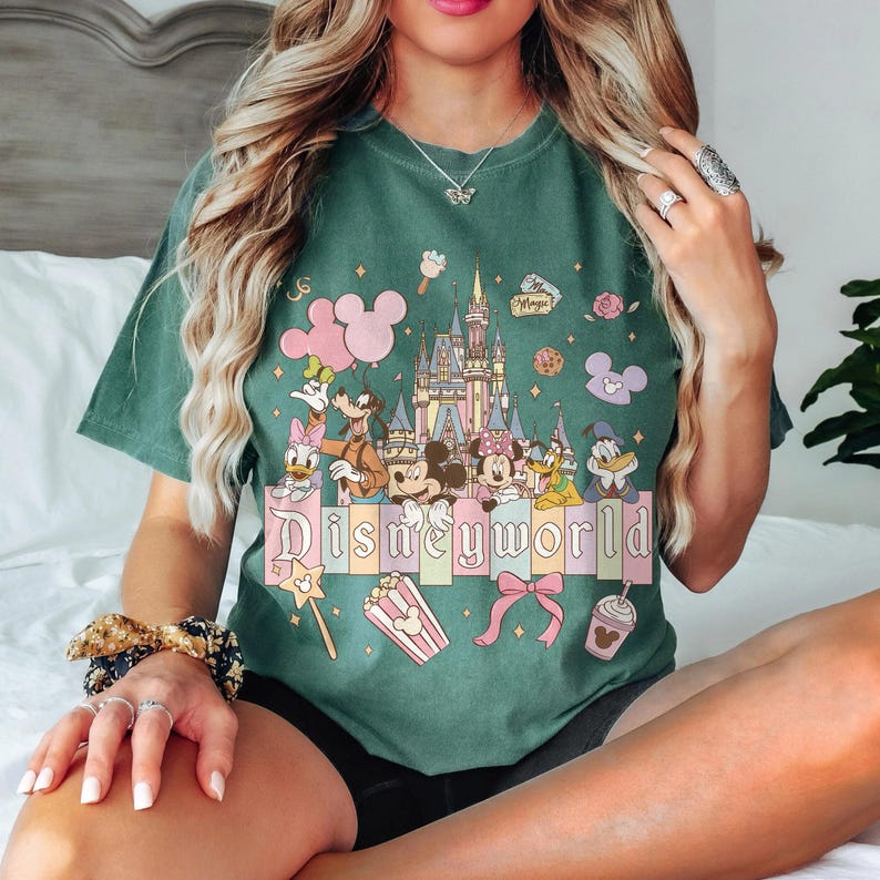 May include: Green t-shirt with a Disney World graphic. The design features Disney characters, a castle, and the text "Disneyworld" in pastel colours. The shirt has a relaxed fit.