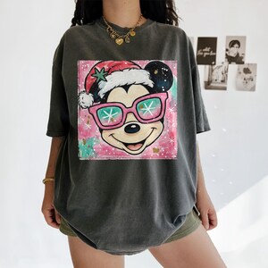 K&ouml;nnte beinhalten: Dunkelgraues T-Shirt mit einem farbenfrohen Aufdruck von Mickey Mouse mit Weihnachtsm&uuml;tze und rosa Sonnenbrille. Die Grafik befindet sich auf einem rosa und t&uuml;rkisfarbenen Hintergrund mit Schneeflocken-Akzenten. Das T-Shirt hat kurze &Auml;rmel.