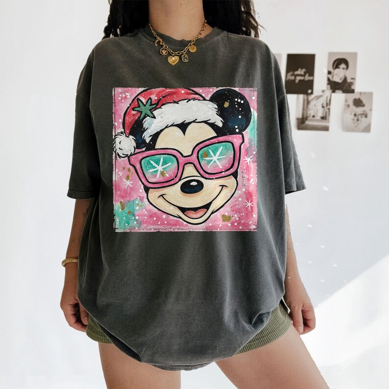 K&ouml;nnte beinhalten: Dunkelgraues T-Shirt mit einem farbenfrohen Aufdruck von Mickey Mouse mit Weihnachtsm&uuml;tze und rosa Sonnenbrille. Die Grafik befindet sich auf einem rosa und t&uuml;rkisfarbenen Hintergrund mit Schneeflocken-Akzenten. Das T-Shirt hat kurze &Auml;rmel.