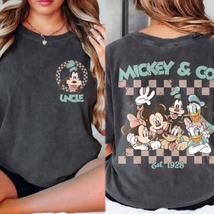 K&ouml;nnte beinhalten: Dunkelgraues T-Shirt mit einem Goofy-Motiv und dem Wort "UNCLE". Auf der R&uuml;ckseite des Shirts sind Micky Maus, Minnie Maus, Donald Duck, Daisy Duck und Goofy mit "MICKEY & CO" und "Est. 1928" abgebildet. Das Design hat einen Retro-Look.