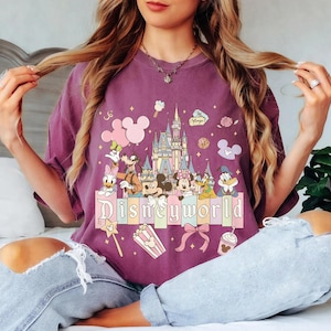 May include: A purple t-shirt with a Disney World design. The graphic features Disney characters, a castle, and the words "Disneyworld" in a colourful font. Additional elements include popcorn, a bow, and a magic wand.