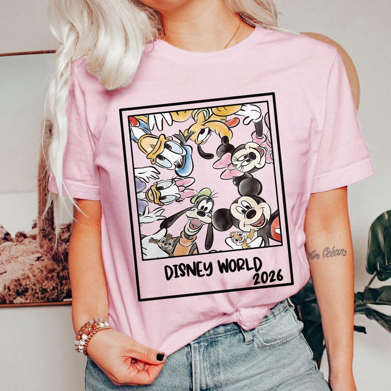 Puede incluir: Camiseta rosa claro con un gr&aacute;fico enmarcado de personajes de Disney, incluyendo Mickey Mouse, Donald Duck y Goofy. El gr&aacute;fico tiene las palabras "Disney World 2026" en fuente negra. La camiseta tiene mangas cortas.