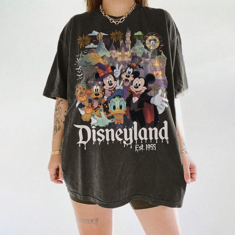 May include: A charcoal gray oversized t-shirt featuring a graphic print of Disney characters in Halloween costumes in front of a castle. The text reads Disneyland Est. 1955 with a dripping effect. The design includes fireworks and bats in the background.