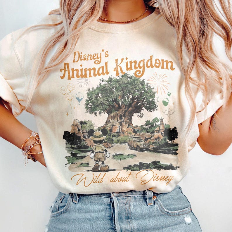 May include: Cream-colored sweatshirt featuring the text "Disney's Animal Kingdom" and "Wild about Disney" in brown script. The graphic depicts a tree, Mickey Mouse in safari attire, and a safari vehicle. The design includes fireworks and hot air balloons.