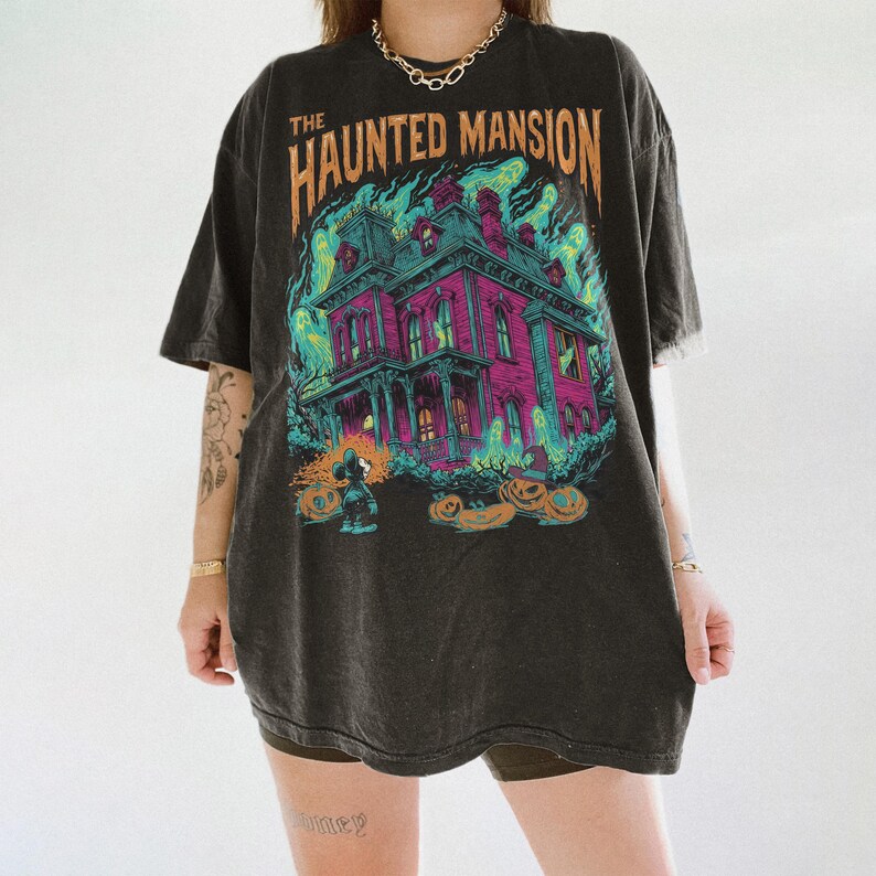 May include: Dark gray t-shirt featuring a colorful graphic of a haunted mansion with the text "THE HAUNTED MANSION". The design includes ghosts, pumpkins, and a cartoon character. The shirt has short sleeves and a relaxed fit.
