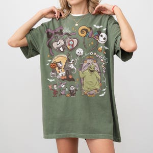 May include: Olive green t-shirt with a colourful graphic design featuring characters from "The Nightmare Before Christmas." The design includes Jack Skellington, Sally, Oogie Boogie, and other iconic imagery. The shirt has a relaxed fit.