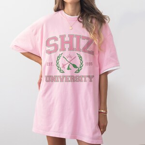 Pu&ograve; includere: T-shirt oversize rosa chiaro con la scritta "SHIZ UNIVERSITY" in stile collegiale. Il design include uno stemma con scope incrociate e un cappello da strega, con "EST. 1995" sotto. La maglietta ha finiture bianche sulle maniche.