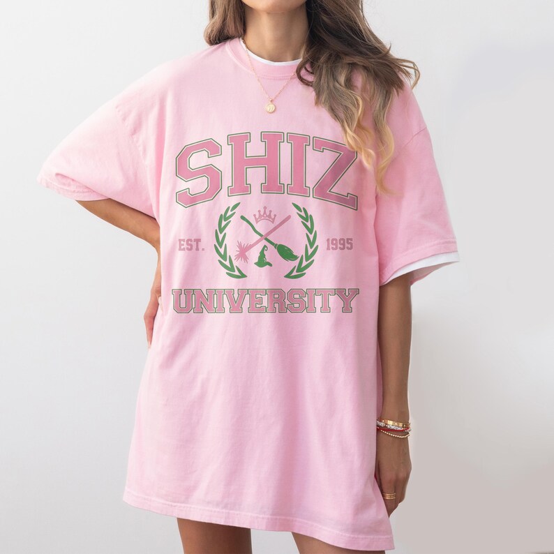 Pu&ograve; includere: T-shirt oversize rosa chiaro con la scritta "SHIZ UNIVERSITY" in stile collegiale. Il design include uno stemma con scope incrociate e un cappello da strega, con "EST. 1995" sotto. La maglietta ha finiture bianche sulle maniche.