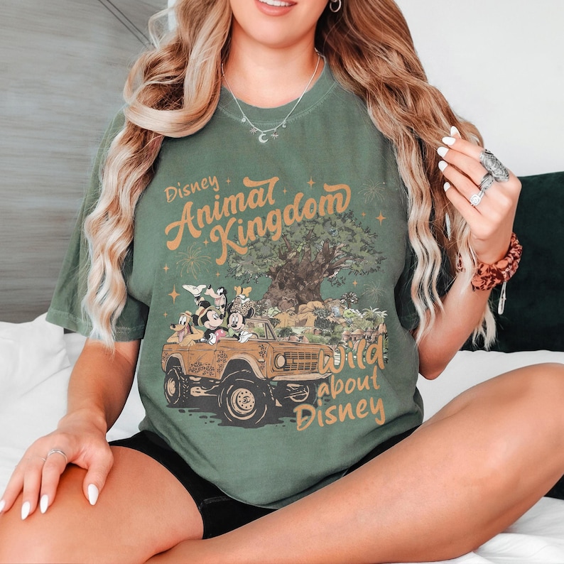 May include: Green t-shirt with the text "Disney Animal Kingdom" and "Wild about Disney". The graphic features Mickey Mouse, Minnie Mouse, and other characters in a safari vehicle with a tree and fireworks.