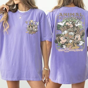 Puede incluir: Una camiseta de color lila con gr&aacute;ficos de personajes de Disney en la parte delantera y trasera. El gr&aacute;fico frontal incluye a Mickey Mouse, el Pato Donald y Goofy. El gr&aacute;fico trasero dice "Animal Kingdom" y "Walt Disney World".