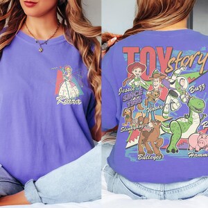 K&ouml;nnte beinhalten: Ein lilafarbenes T-Shirt mit einem Front-Grafik einer Prinzessin und dem Namen "Kiara". Auf der R&uuml;ckseite des Shirts befindet sich eine "Toy Story"-Grafik mit Figuren wie Woody, Buzz und Rex. Der Text "Toy Story" ist rot.