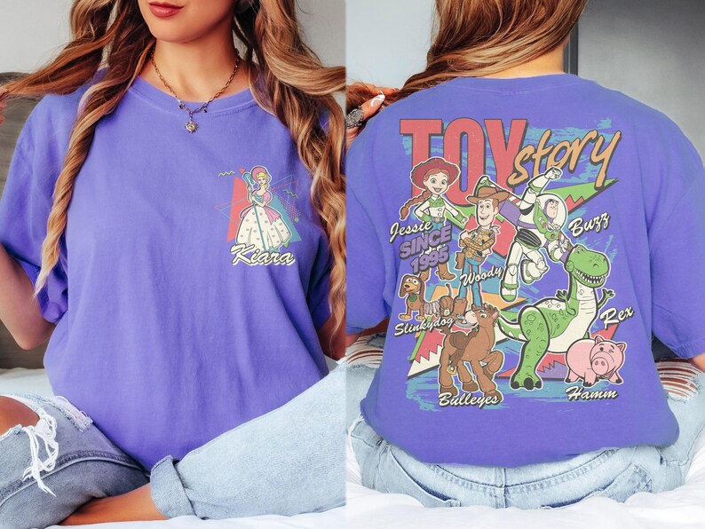 K&ouml;nnte beinhalten: Ein lilafarbenes T-Shirt mit einem Front-Grafik einer Prinzessin und dem Namen "Kiara". Auf der R&uuml;ckseite des Shirts befindet sich eine "Toy Story"-Grafik mit Figuren wie Woody, Buzz und Rex. Der Text "Toy Story" ist rot.