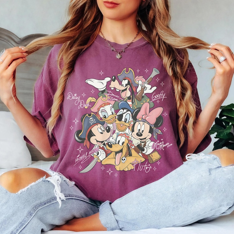 May include: A purple t-shirt featuring a graphic of Disney characters dressed as pirates. The characters include Mickey Mouse, Minnie Mouse, Donald Duck, Daisy Duck, Goofy, and Pluto. The shirt has the words "Mickey Mouse" and "Minnie Mouse" printed on it.