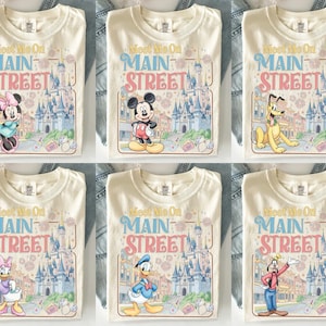K&ouml;nnte beinhalten: Cremefarbene T-Shirts mit dem Text "Meet Me On Main Street" und verschiedenen Zeichentrickfiguren. Die Shirts zeigen Minnie Mouse, Mickey Mouse, Pluto, Daisy Duck, Donald Duck und Goofy, mit einer Schlosskulisse.