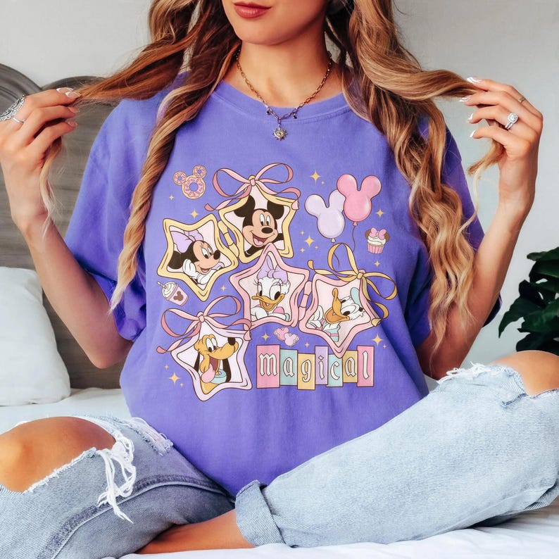 May include: A lilac-colored t-shirt featuring a graphic design of Disney characters inside star-shaped frames. The design includes Mickey Mouse, Minnie Mouse, Donald Duck, Daisy Duck, and Pluto, with the word "magical" below.