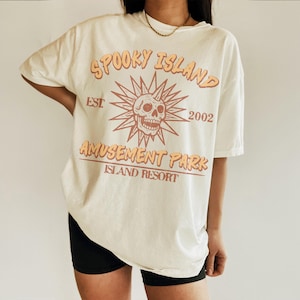 May include: Cream-colored t-shirt with a distressed graphic. The design features the text "Spooky Island Amusement Park Island Resort" with a skull and sunburst illustration. The shirt has a vintage aesthetic.
