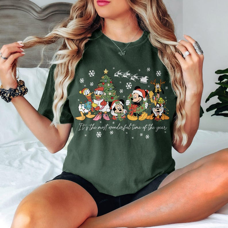 May include: Dark green t-shirt featuring a festive graphic of Disney characters, including Mickey and Minnie Mouse, Donald Duck, and Pluto, with a Christmas tree and snowflakes. The text "It's the most wonderful time of the year" is printed below the characters.