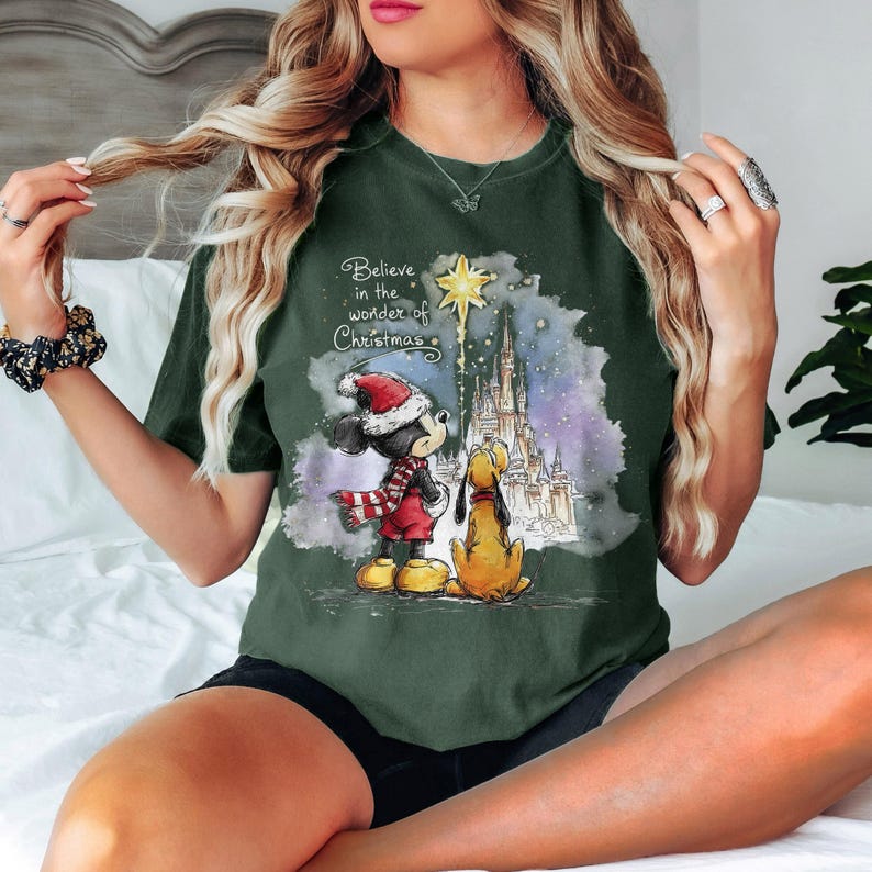 May include: Dark green t-shirt featuring a Christmas-themed graphic. The graphic includes Mickey Mouse in a Santa hat and Pluto, with a castle and the text "Believe in the wonder of Christmas".