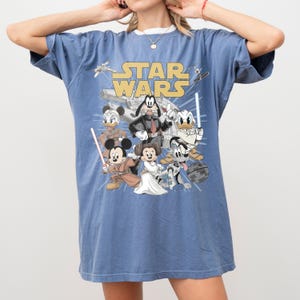 May include: Blue t-shirt featuring a graphic of Star Wars characters, including Mickey Mouse as Luke Skywalker, Goofy as Darth Vader, and Donald Duck. The words "STAR WARS" are printed in yellow.