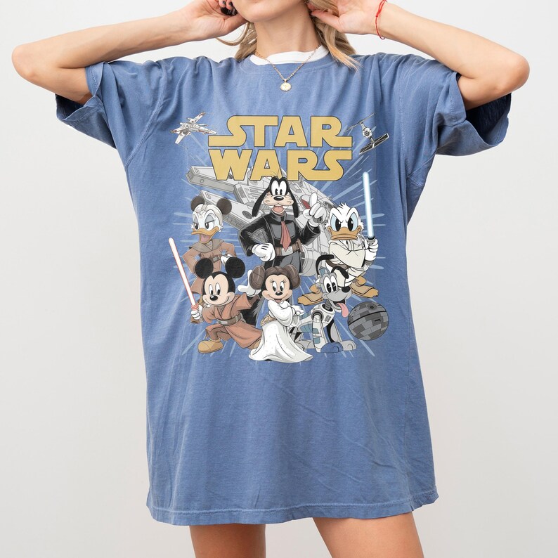 May include: Blue t-shirt featuring a graphic of Star Wars characters, including Mickey Mouse as Luke Skywalker, Goofy as Darth Vader, and Donald Duck. The words "STAR WARS" are printed in yellow.