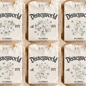 May include: Cream-coloured t-shirts with the text "Disneyworld" above the text "est. 1971 FLORIDA". Each shirt features a different cartoon character decorated with Christmas lights. The shirts are folded and arranged on a woven surface.