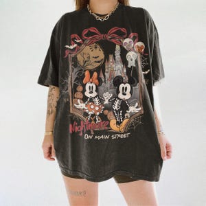 May include: Black t-shirt featuring a Halloween-themed graphic. The design includes Mickey and Minnie Mouse in skeleton costumes, a castle, and the words "Nightmare on Main Street." The shirt has a vintage, worn look.
