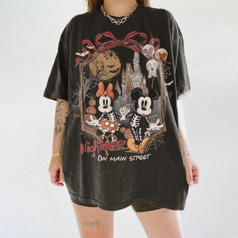 May include: Black t-shirt featuring a Halloween-themed graphic. The design includes Mickey and Minnie Mouse in skeleton costumes, a castle, and the words "Nightmare on Main Street." The shirt has a vintage, worn look.