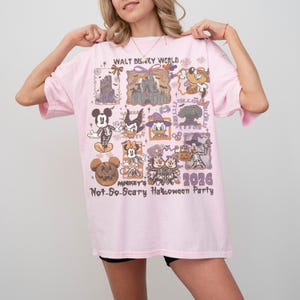 May include: A light pink t-shirt featuring a colorful graphic design with various Disney characters and the text "WALT DISNEY WORLD" and "Mickey's Not-So-Scary Halloween Party 2026". The shirt has short sleeves.