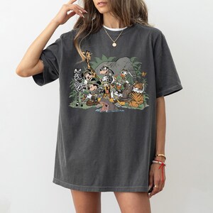 K&ouml;nnte beinhalten: Dunkelgraues T-Shirt mit einer farbenfrohen Cartoon-Illustration von Disney-Figuren als Safari-Entdecker, darunter ein Zebra, eine Giraffe, ein Elefant und ein Tiger. Micky Maus, Donald Duck und Goofy sind abgebildet. Das Shirt hat eine lockere Passform.