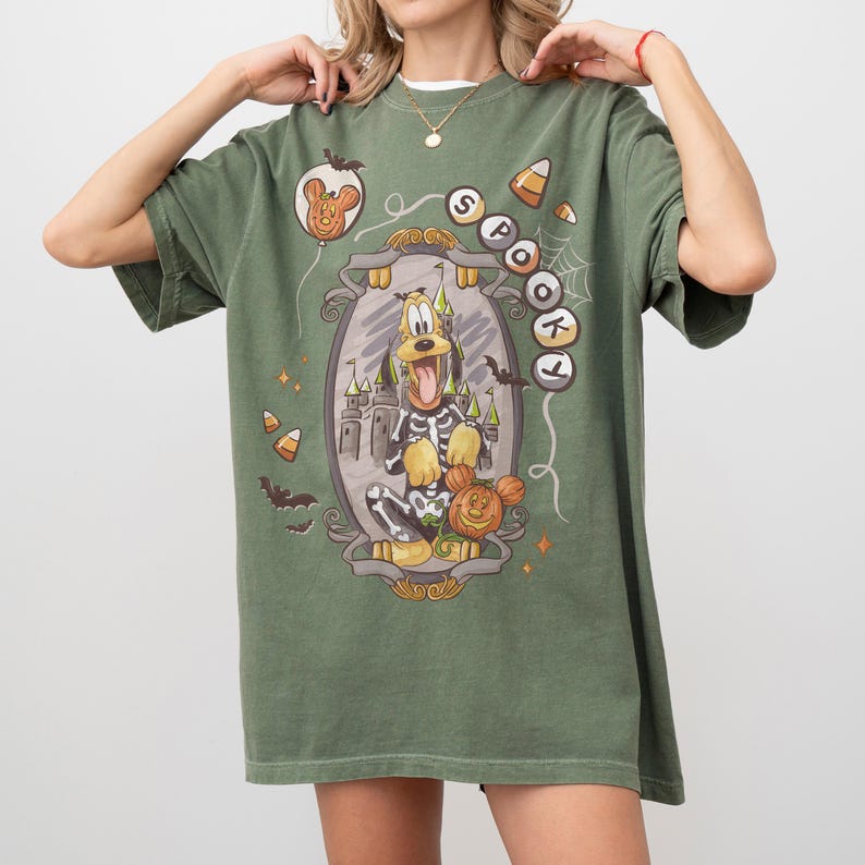 May include: Olive green t-shirt featuring a Halloween-themed graphic. The graphic includes a cartoon dog in a skeleton costume, a pumpkin, and the word "Spooky". Additional elements include bats, candy corn, and a castle.