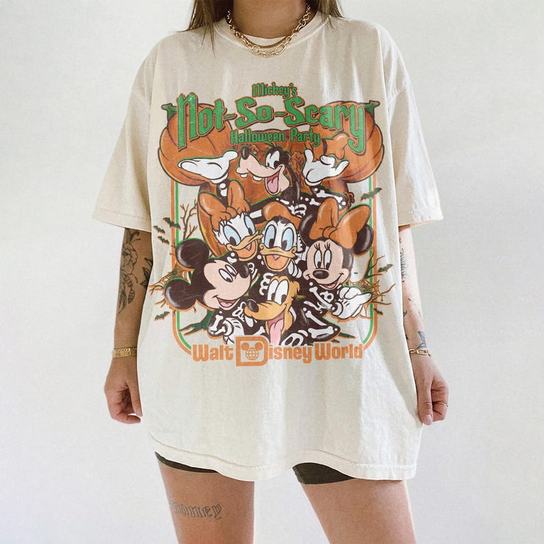 May include: An off-white short-sleeve t-shirt featuring a graphic print of Mickey Mouse, Minnie Mouse, Donald Duck, Daisy Duck, and Goofy wearing skeleton costumes. The text reads Mickey's Not-So-Scary Halloween Party and Walt Disney World.
