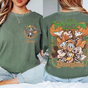 May include: A sage green t-shirt featuring a small graphic on the front chest of Goofy in a skeleton costume with the word Uncle. The back displays a large graphic of Mickey, Minnie, Donald, Daisy, and Goofy in skeleton costumes with the text Mickey's Trick-or-Treat Halloween Party Disneyland.