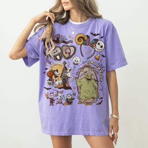 May include: A lilac t-shirt with a vibrant graphic design featuring characters and elements from "The Nightmare Before Christmas." The design includes Jack Skellington, Sally, and Oogie Boogie, along with other Halloween-themed imagery.